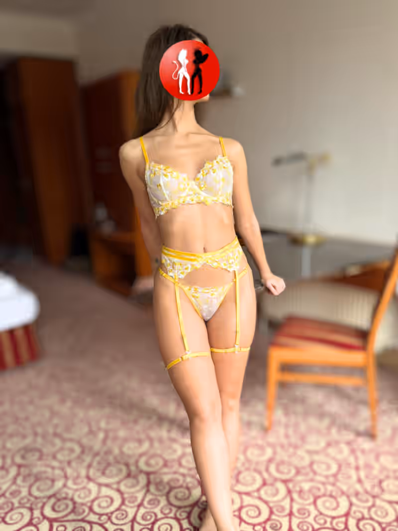 Escort Celina aus Leipzig steht im Hotelzimmer und trägt ein zartes, gelbes Dessous-Set mit Strapsen. Ihre Haltung ist leicht verspielt, ihr Körper wirkt schlank und anziehend. Eine junge Frau, die weiß, wie man Lust mit Leichtigkeit verbindet.