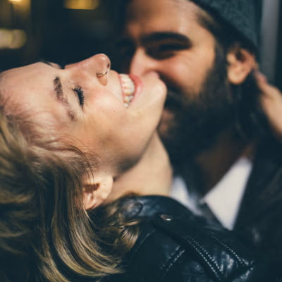 Ein verliebtes Paar lacht innig miteinander. Die Frau lehnt sich mit geschlossenen Augen und strahlendem Lächeln zurück, während der Mann sie zärtlich an sich zieht. Beide wirken entspannt und glücklich – eine Momentaufnahme voller Nähe, Leichtigkeit und Freude. Die Frau trägt eine schwarze Lederjacke, der Mann eine Mütze und einen Bart. Die Szene ist leicht unscharf im Hintergrund und strahlt eine warme, verspielte Intimität aus.