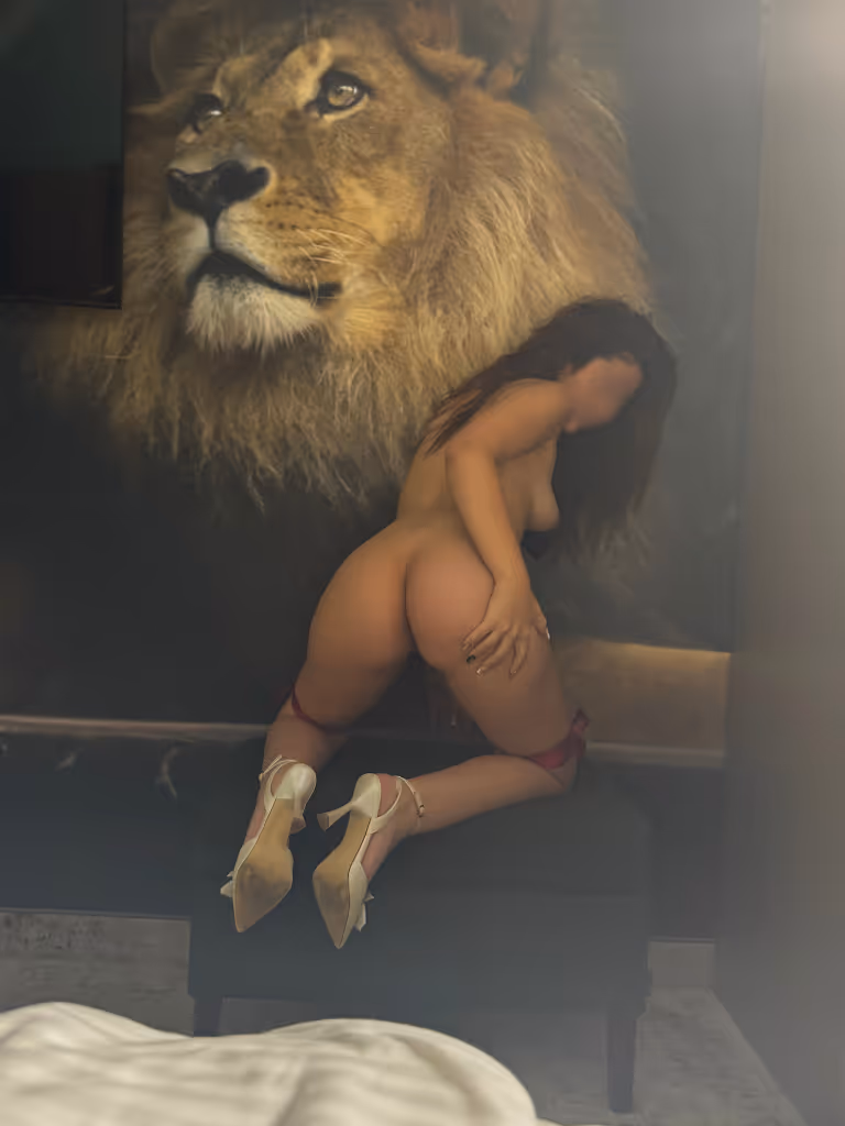 Mila posiert sinnlich nackt auf einem Hocker, während im Hintergrund ein imposantes Löwenbild ihre wilde und selbstbewusste Ausstrahlung unterstreicht.