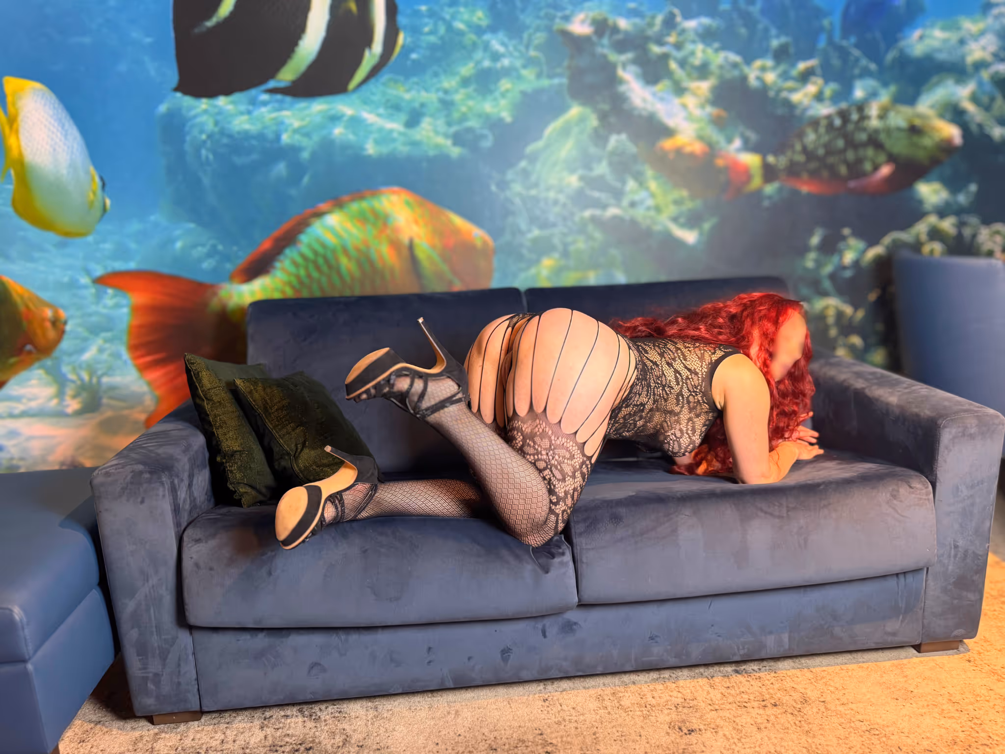 Romy rothaarige Escort Lady in Leipzig kniet auf dem roten Sofa Rückansicht. Glänzendes rotes Latex Minikleid betont ihre Kurven lange rote Haare fallen über den Rücken schwarze High Heels sinnliche und einladende Pose vor Aquarium Wandbild Luxus Hotel Leipzig.