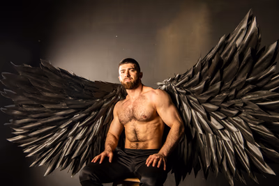 Ein muskulöser, oberkörperfreier Mann sitzt auf einem Hocker vor einem dunklen Hintergrund. Er trägt schwarze Hosen und blickt mit ruhigem, selbstbewusstem Ausdruck in die Kamera. Besonders auffällig sind die großen, kunstvoll gearbeiteten schwarzen Flügel aus einem federartigen Material, die sich weit nach beiden Seiten ausbreiten – wie bei einem gefallenen Engel oder einem dunklen Schutzgeist. Das Licht hebt die Struktur seiner Muskeln und der Flügel eindrucksvoll hervor. Er könnte ein Escort Lieblingskunde sein.