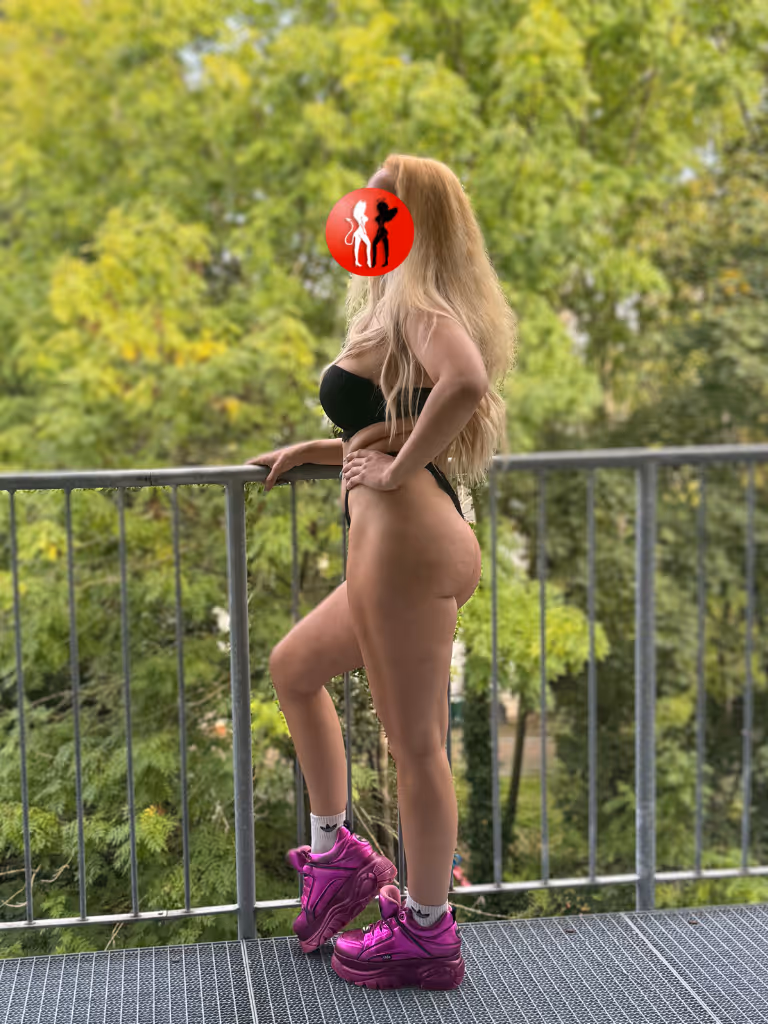 Escort Sophia aus Leipzig steht selbstbewusst auf einem Balkon, nur in schwarzem Dessous und pinken Sneakers. Ihr knackiger Po, die langen blonden Haare und die Pose mit Hüftschwung versprühen pure Lebenslust – sexy, frech und voller Energie.