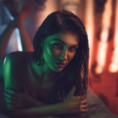 Eine sinnliche Frau mit nassen Haaren liegt auf einem Bett und blickt direkt in die Kamera. Ihr Körper ist nur leicht bedeckt, während dramatisches, farbiges Licht in Grün- und Rottönen ihr Gesicht und ihre Silhouette akzentuiert. Die Atmosphäre wirkt geheimnisvoll, verführerisch und leicht verrucht, unterstützt durch die weichen, unscharfen Lichtquellen im Hintergrund. Ihr Blick ist intensiv, herausfordernd und zugleich spielerisch – eine Einladung an die Fantasie.