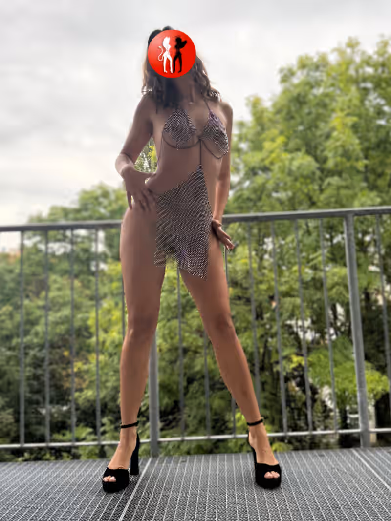 Escort Vivienne aus Leipzig steht halbnackt auf einem Balkon, nur in einem transparenten Glitzer-Outfit, das kaum etwas verdeckt. Ihre langen Beine, der freie Blick auf ihre Brüste und die selbstbewusste Pose in High Heels machen das Bild zu einem echten erotischen Statement – sexy, schamlos und provokant.