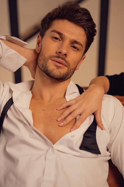 Auf dem Bild ist ein attraktiver Mann mit leicht geöffnetem, weißen Hemd und schwarzer Fliege zu sehen, die locker um seinen Hals hängt. Er schaut selbstbewusst und sinnlich in die Kamera. Seine Haltung ist entspannt – ein Arm liegt im Nacken, während eine weibliche Hand zärtlich über seine Brust streicht. Die Szene wirkt intim, verführerisch und lädt zum Kopfkino ein – perfekt passend zum Thema „Bin ich gut im Bett?“.