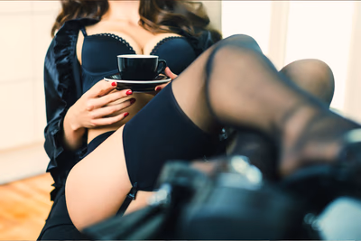 Eine Frau in schwarzen Dessous sitzt zurechtgemacht auf einem Stuhl, hält einen Kaffee in der Hand und scheint auf etwas zu warten