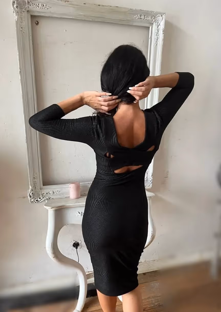 Anna steht mit dem Rücken zur Kamera in einem figurbetonten, schwarzen Kleid mit raffinierten Cut-Outs. Sie steckt sich gerade die Haare hoch, was ihren eleganten Nacken freilegt. Im Hintergrund befindet sich ein dekorativer, weißer Vintage-Spiegel auf einer Konsole. Das Bild strahlt Stil, Klasse und eine subtile Spannung aus.

