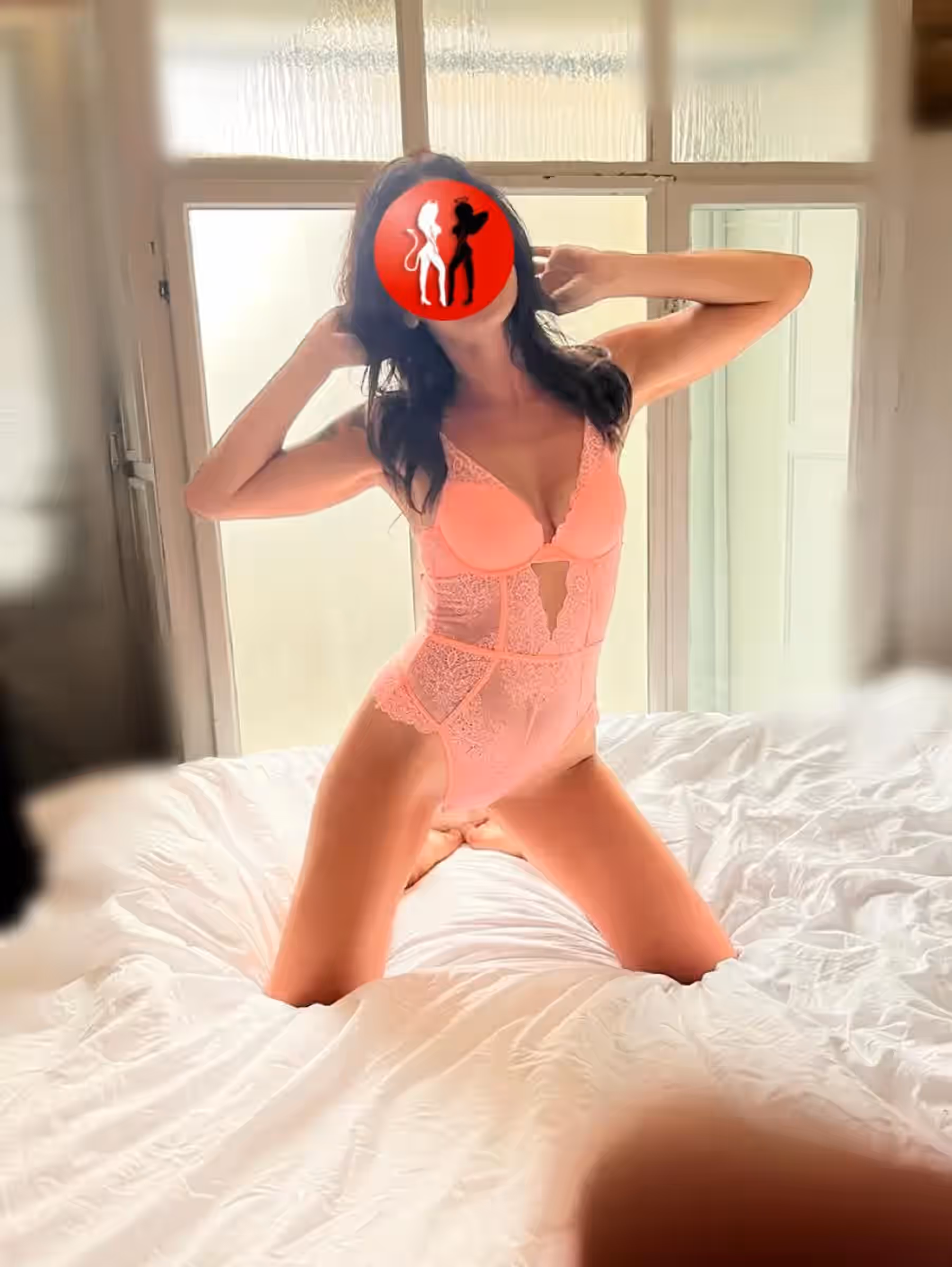 In dieser Szene kniet Anna auf einem weißen Bett vor einem lichtdurchfluteten Fenster. Sie trägt einen rosafarbenen Spitzenbody mit tiefem Ausschnitt. Ihre Pose ist lebendig, ihre Hände berühren das Haar, was dem Bild Bewegung und Sinnlichkeit verleiht. Die weiche Lichtführung sorgt für eine warme, intime Atmosphäre.