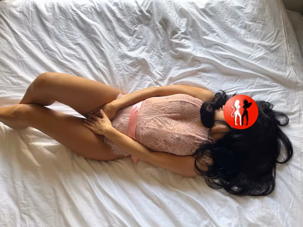 Julia liegt ausgestreckt auf dem Rücken auf einem weißen Bett. Sie trägt einen zart rosafarbenen Spitzenbody mit breitem Satinschleifenband um die Taille. Ihre dunklen Haare fallen locker zur Seite, die Pose ist ruhig, feminin und wirkt verträumt. Die weiche Bettwäsche und das Tageslicht verleihen dem Bild eine entspannte, intime Stimmung.