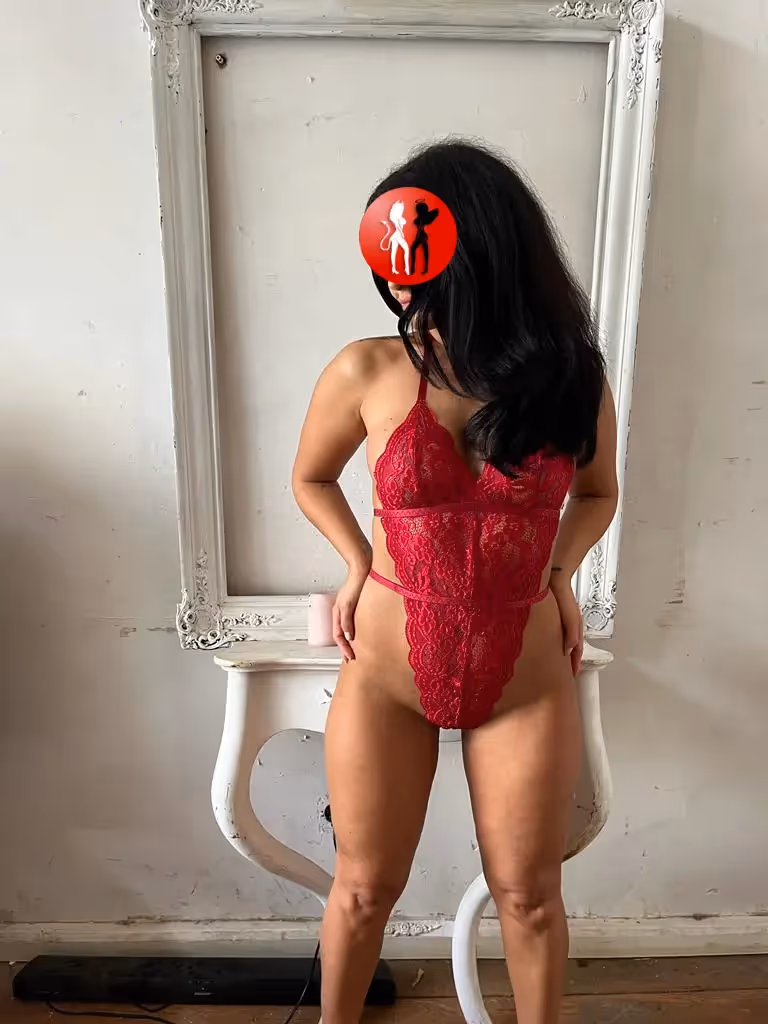 Julia steht aufrecht vor einem verzierten weißen Rahmen, der als stilistisches Element die Szenerie abrundet. Sie trägt einen roten Spitzenbody, der ihre Taille und Silhouette betont. Ihre dunklen, leicht gewellten Haare fallen über eine Schulter. Der Gesamteindruck ist feminin, stark und zugleich verspielt.