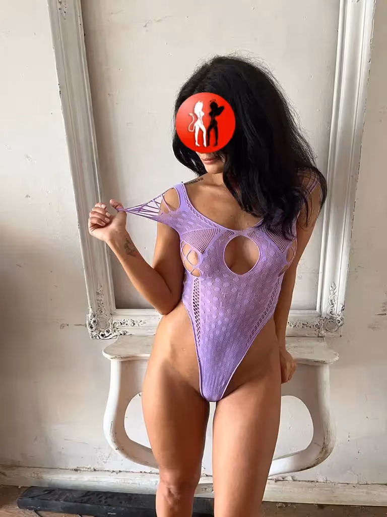 Vor einem barocken, weißen Spiegelrahmen steht Julia in einem auffälligen lilafarbenen Netzbody mit ovalen Öffnungen im Brust- und Schulterbereich. Ihre Pose ist verspielt: Sie zieht neckisch am Träger ihres Bodys, während ihr Blick verborgen bleibt. Die Szene wirkt kokett und zugleich kunstvoll in Szene gesetzt.

