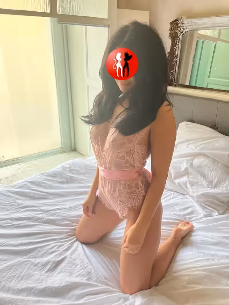 Julia kniet auf einem weiß bezogenen Bett in einem hellen, stilvollen Schlafzimmer. Sie trägt denselben rosa Spitzenbody mit Satinband, der ihre schlanke Figur unterstreicht. Im Hintergrund befindet sich ein verzierter Spiegelrahmen sowie ein lichtdurchflutetes Fenster. Ihre Pose ist aufrecht, natürlich und wirkt offen, aber dezent.