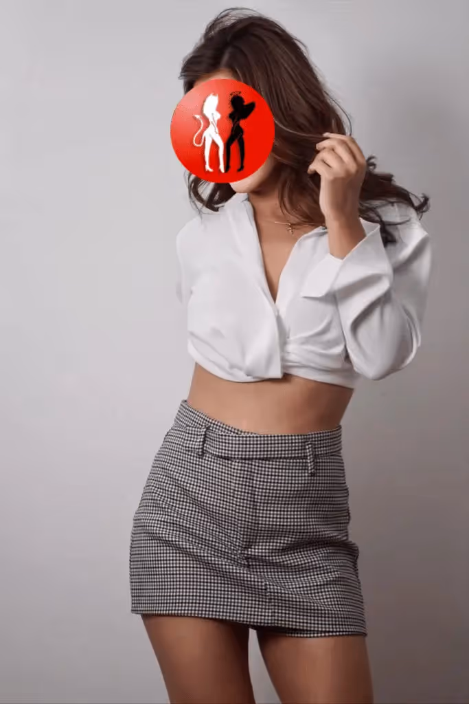 Valeria trägt eine schlichte, weiße Bluse, die vorne locker geknotet ist, und kombiniert dazu einen kurzen, karierten Minirock. Sie steht vor neutralem Hintergrund, eine Hand spielt mit ihrem Haar. Der Look ist modern, feminin und erinnert an einen selbstbewussten Office-Style mit verführerischem Understatement.