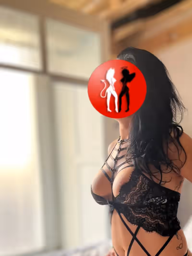 Vanessa steht in einem lichtdurchfluteten Raum, gekleidet in dieselbe kunstvolle schwarze Spitzen-Lingerie mit Bondage-Akzenten. Der Bildfokus liegt auf ihrem Oberkörper und ihrer Silhouette. Ihr Gesicht ist anonymisiert, aber ihre Pose vermittelt Präsenz, Selbstsicherheit und feminine Eleganz. Das diffuse Tageslicht sorgt für eine warme, weiche Lichtstimmung.