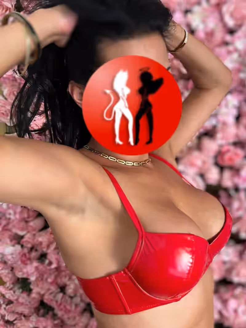 In diesem lebendigen Bild steht Vanessa vor einer opulenten Wand aus rosa Blüten. Sie trägt roten Lack-BH, goldenen Halsschmuck und hebt beide Arme hoch, wodurch ihr Haar locker nach hinten fällt. Die Farbkombination aus Rot, Rosa und Gold erzeugt eine auffällige, aber harmonische Bildwirkung – selbstbewusst, feminin, kraftvoll.