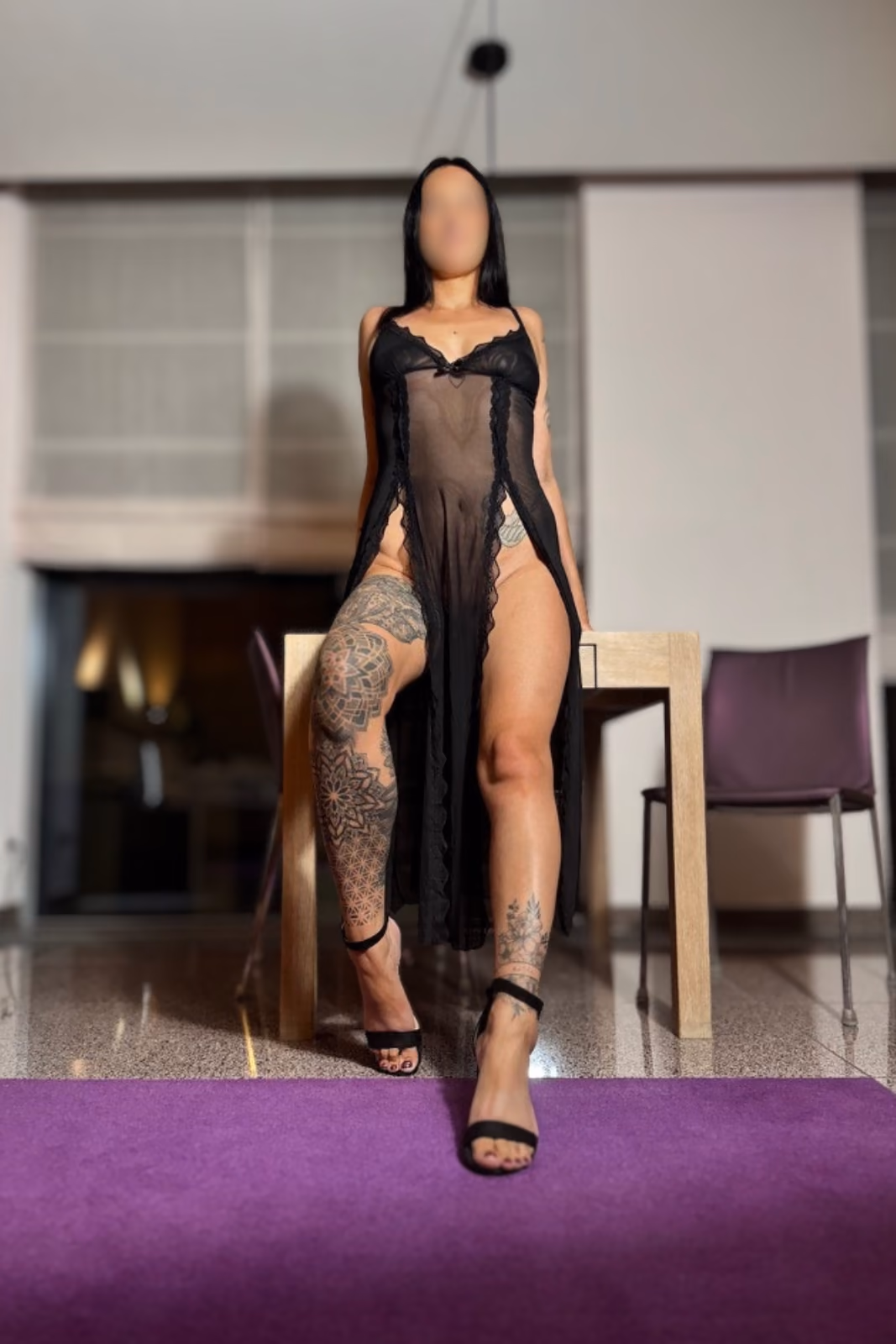 Sophie steht in einem schwarzen Negligé und Sandaletten vor einem Holztisch; ihre langen Beine und Beintattoos sind sichtbar.